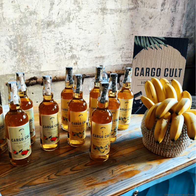 Cargo Cult Rum Moodbild - Viele Flaschen stehen nebeneinander und neben einem Korb mit Bananen auf einem Tisch